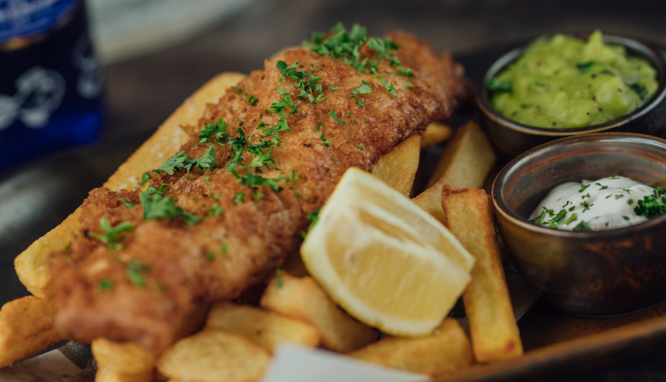 Plate of battered fish and chunky chips with lemon wedge, mushy peas, and tartar sauce.